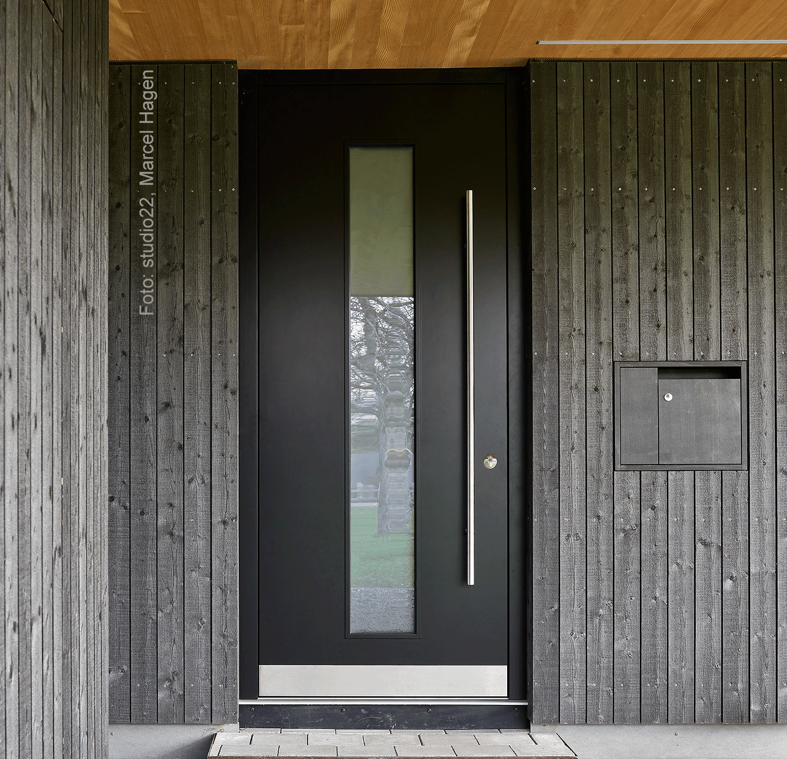 Modern black front door with vertical glass panel and sleek metal handle, framed by dark wood-paneled walls and a mailbox to the right.
