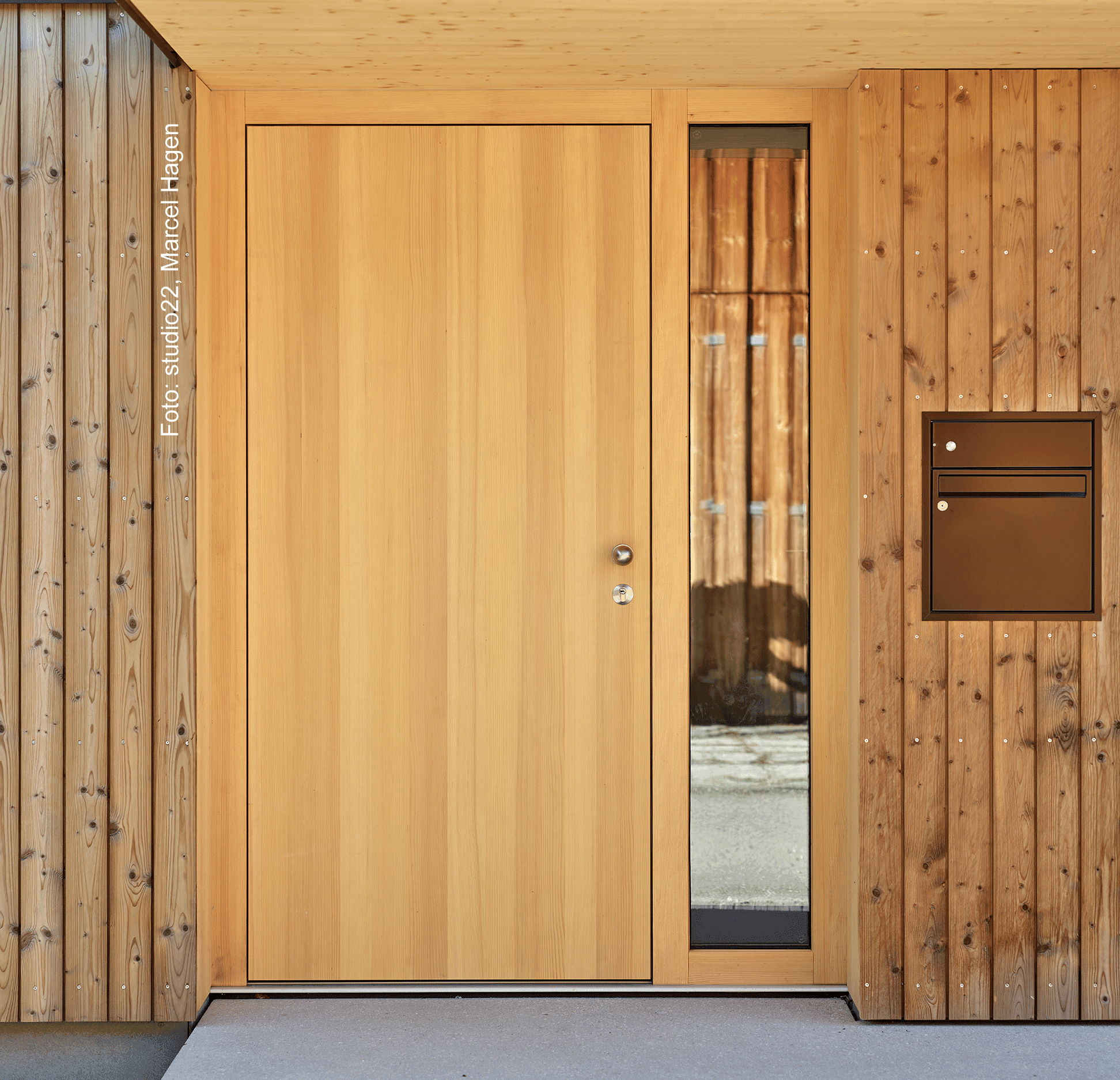 Moderne Holztür mit vertikaler Maserung, seitlicher Glasscheibe und braunem Briefkasten, eingefasst in eine helle Holzfassade.