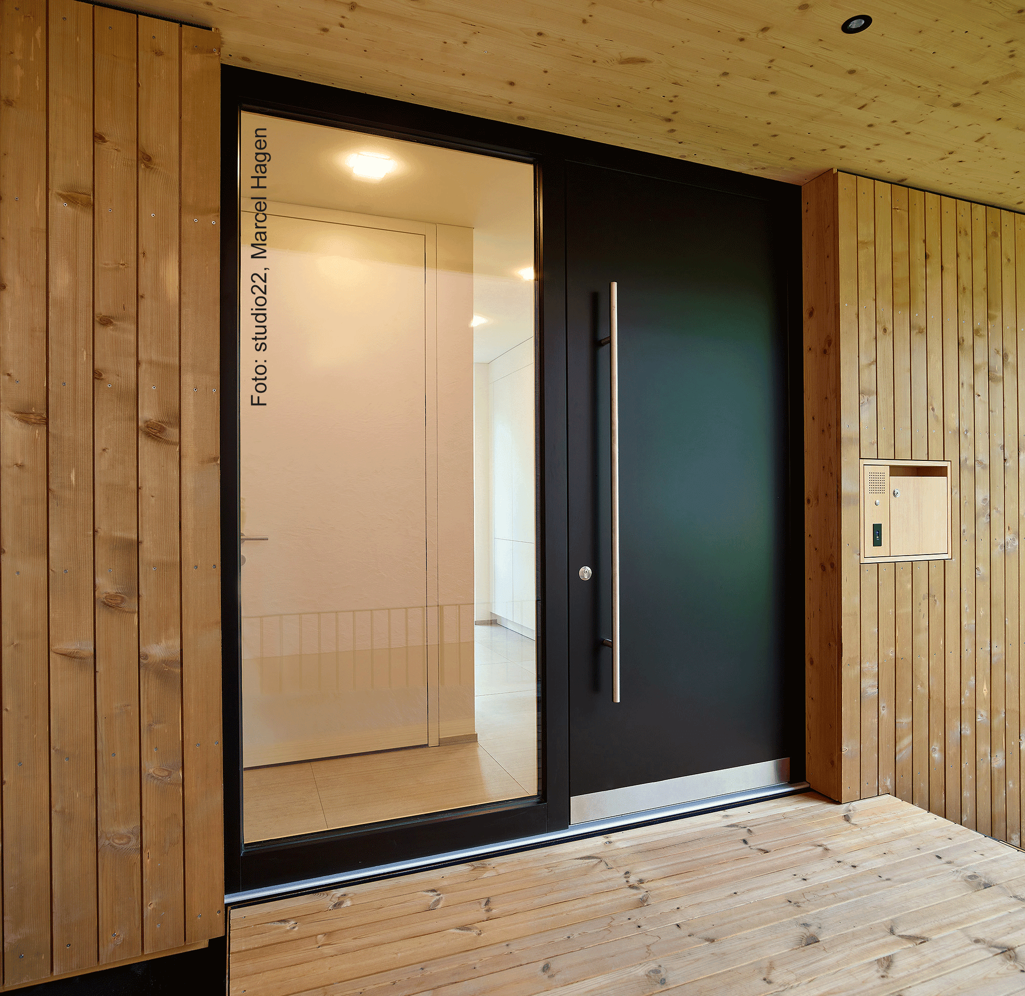Modern wooden entryway with a large glass door, sleek handle, and lighted interior. Wood paneling and a small mailbox are on the right.